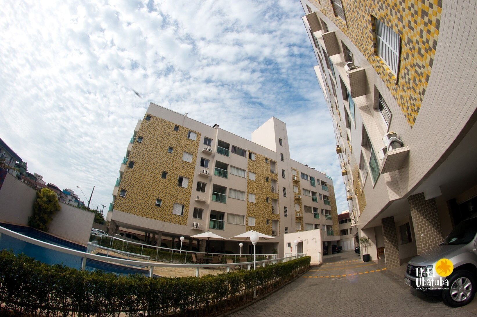 Un edificio de varios pisos con fachada decorativa, un espacio exterior cubierto con asientos y un área de estacionamiento con un automóvil.