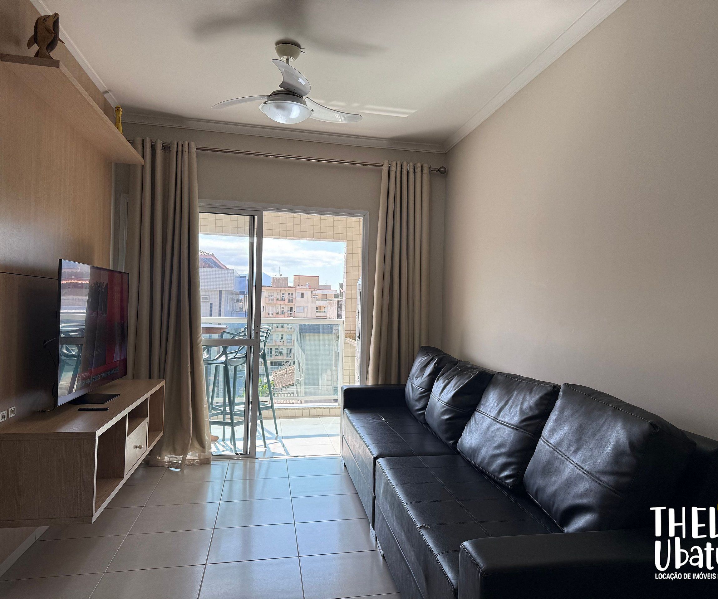 Una sala de estar moderna con un sofá de cuero, un televisor de pantalla plana sobre un mueble de madera, un ventilador de techo y un balcón con vista a edificios.
