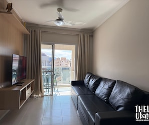 Un salon moderne avec un canapé en cuir, un téléviseur à écran plat posé sur un meuble en bois, un ventilateur de plafond et un balcon offrant une vue sur les bâtiments.