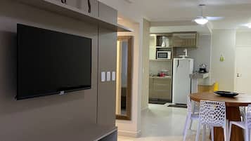 Una sala de estar moderna con un televisor de pantalla plana montado en la pared, una zona de comedor con una mesa de madera y sillas blancas, y una cocina con microondas y refrigerador.