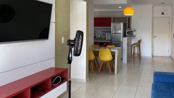 A modern living room with a flat-screen TV, a standing fan, and a dining area with yellow chairs.