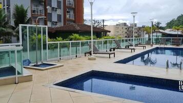 Área de piscina no terraço, com guarda-corpos de vidro, espreguiçadeiras e vista para edifícios ao redor.
