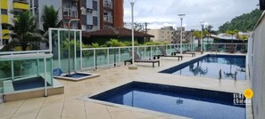 Área de piscina no terraço, com guarda-corpos de vidro, espreguiçadeiras e vista para edifícios ao redor.