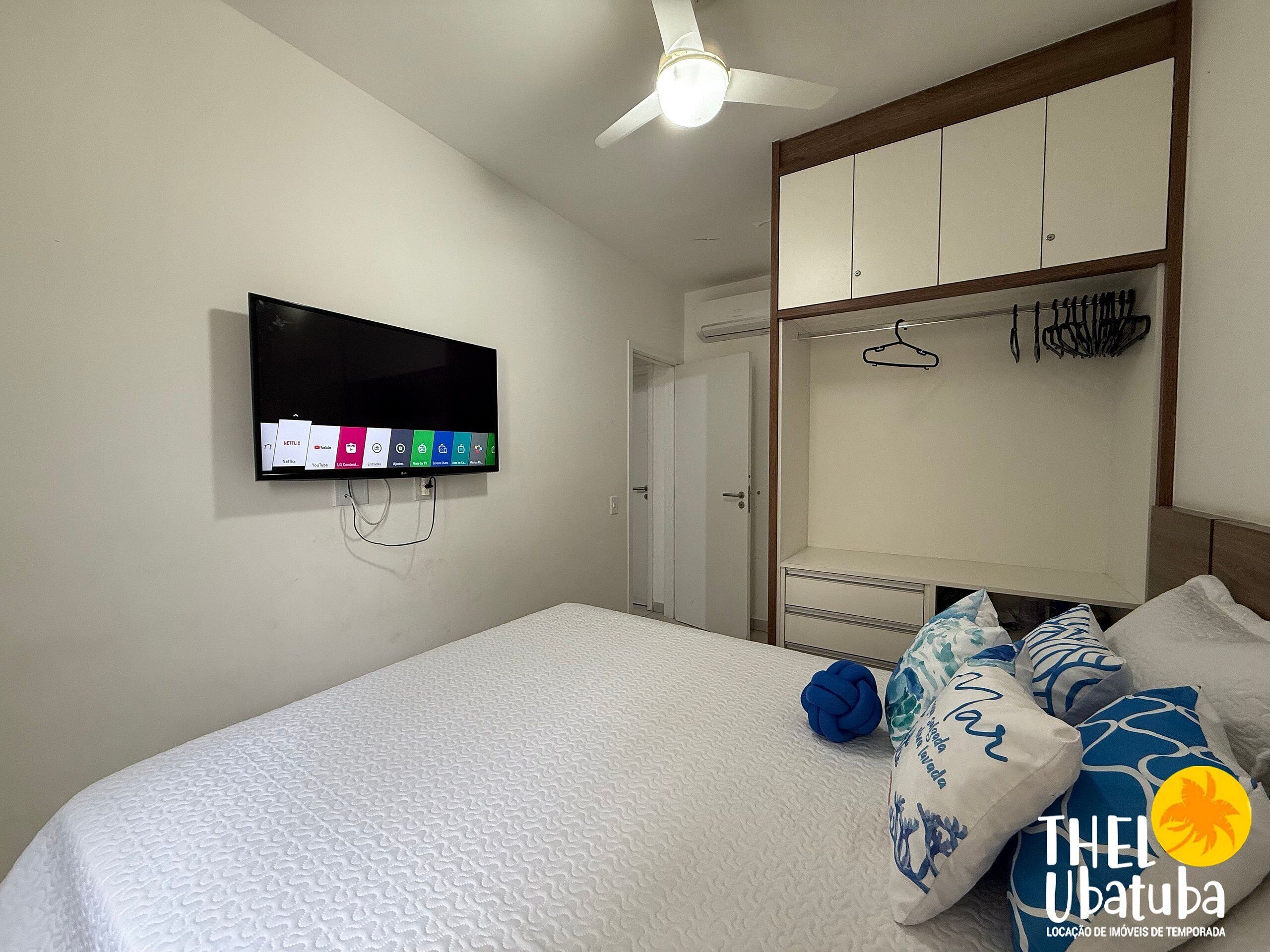 Quarto com cama, televisão na parede, guarda-roupa e ventoinha no teto.