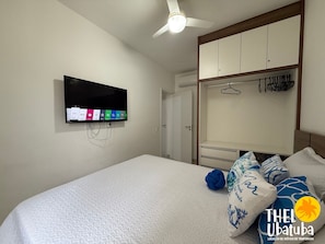 Quarto com cama, televisão na parede, guarda-roupa e ventoinha no teto.