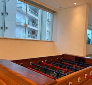 A room with a foosball table, a window with a view of a building, and a billiards table.