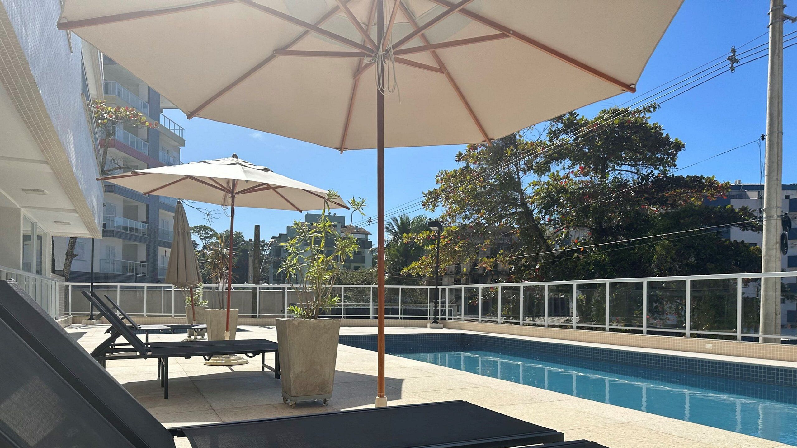 Zona de piscina con sillas de descanso y sombrillas, rodeada por una cerca blanca y edificios al fondo.