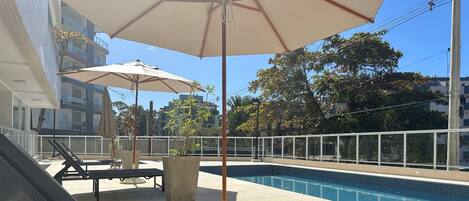 Zona de piscina con sillas de descanso y sombrillas, rodeada por una cerca blanca y edificios al fondo.