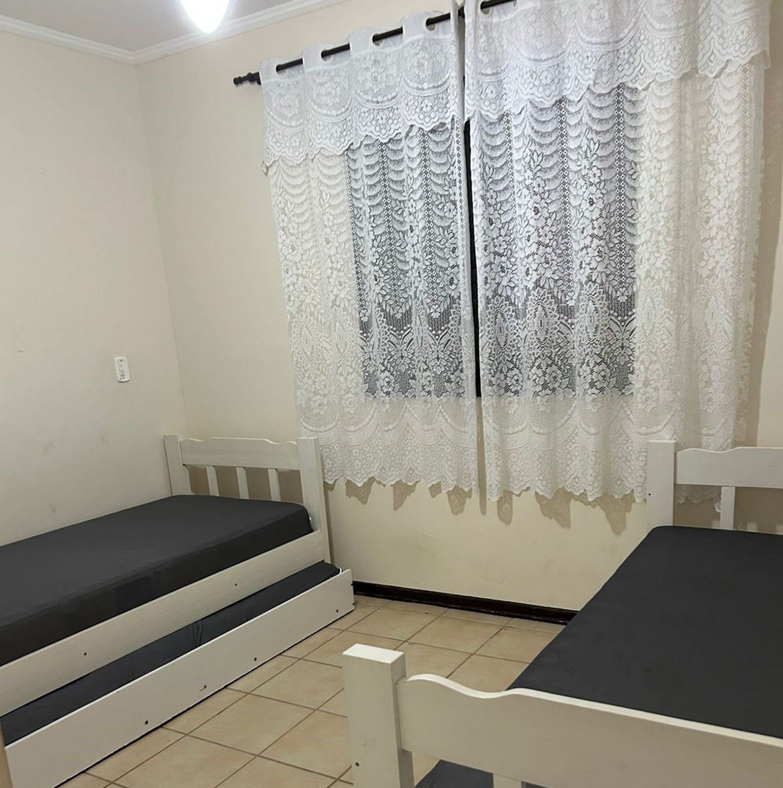 Quarto pequeno e limpo com uma cama de solteiro, uma mesa e uma janela com cortinas de renda.