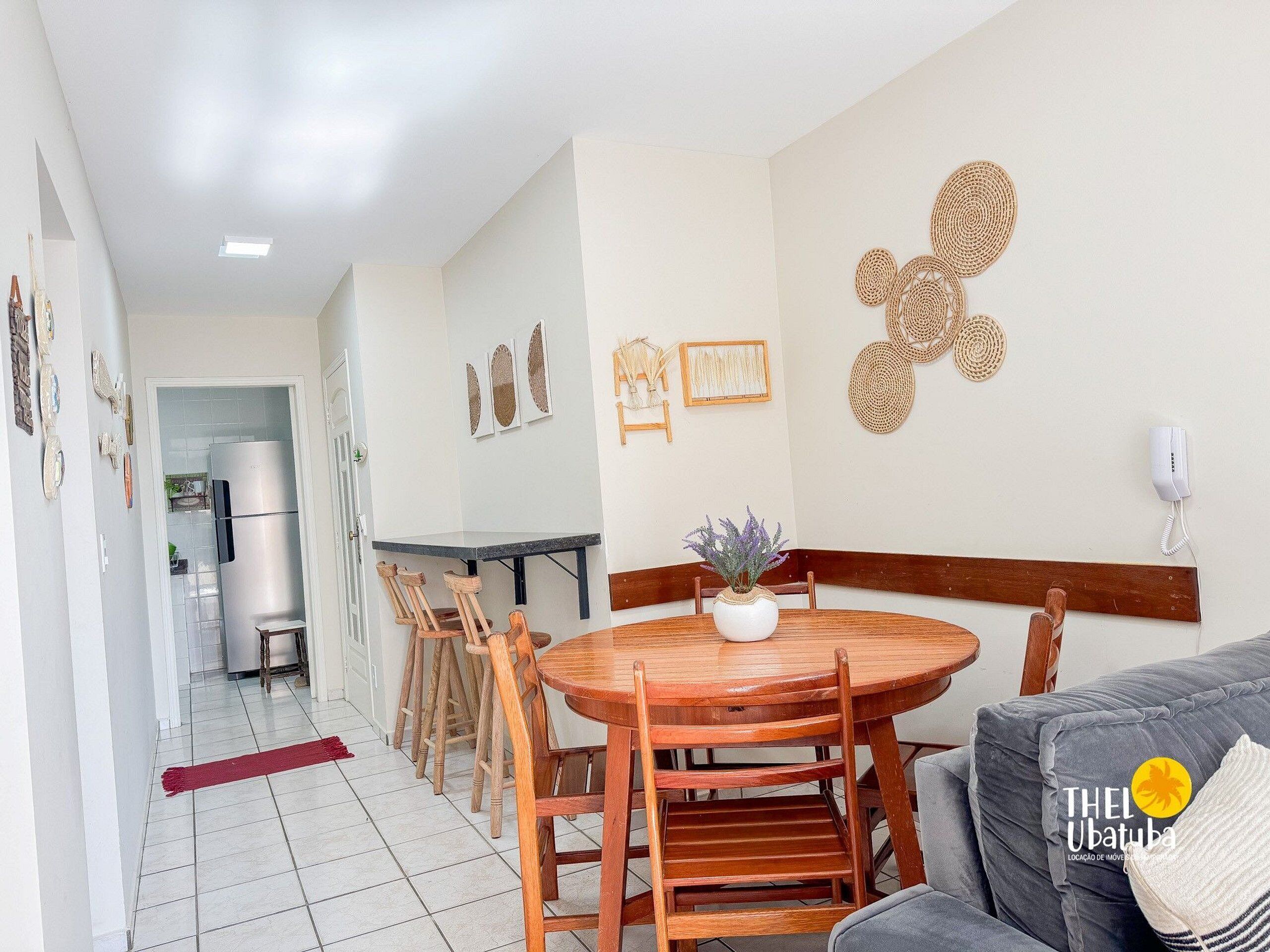 A dining area with a round wooden table, chairs, and a grey sofa. There is a kitchen in the background with a refrigerator and a sink.
