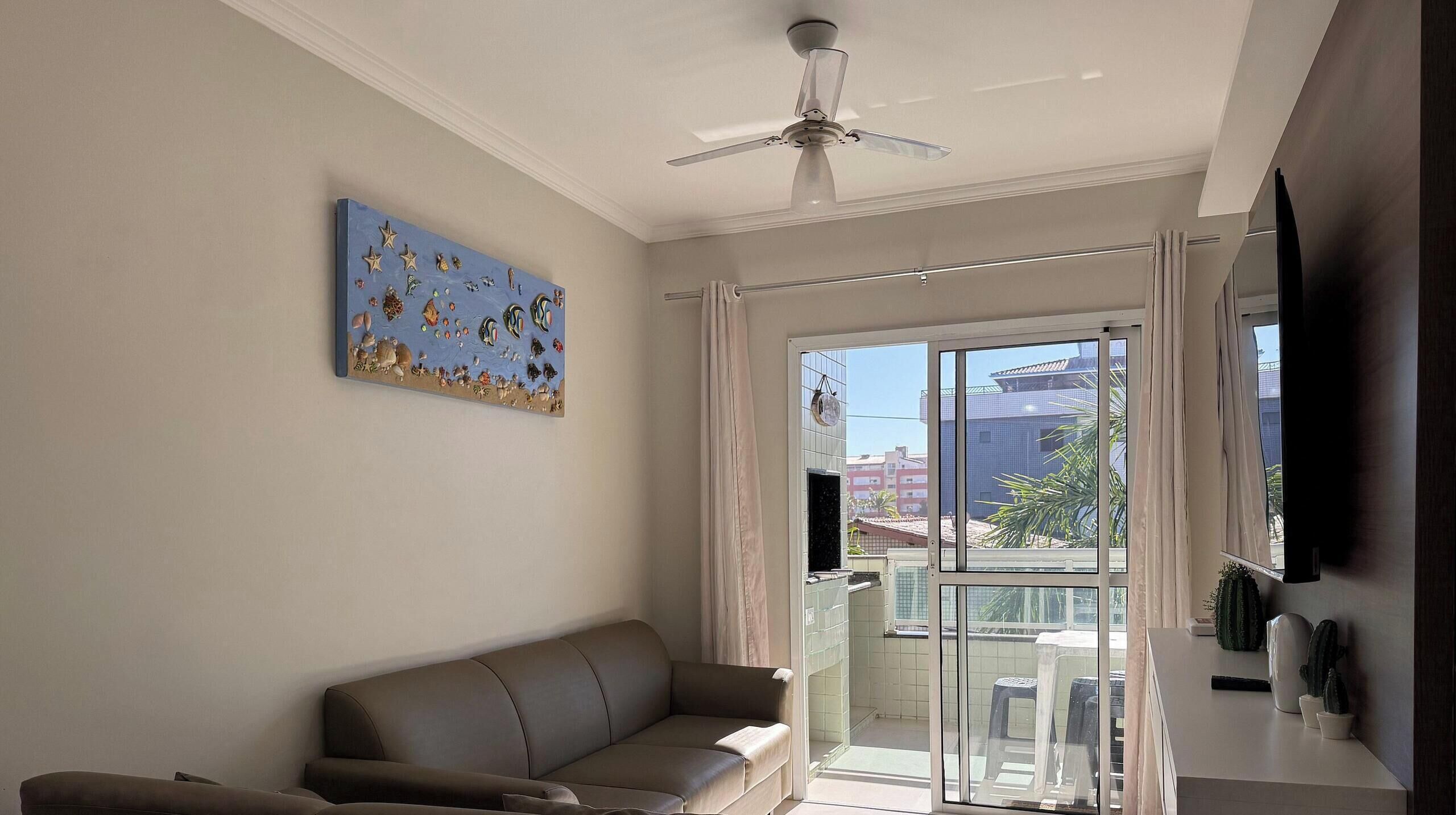 A living room with a sofa, a ceiling fan, a large window with curtains, and a wall-mounted TV.