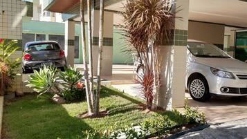 A parking area with two cars, a building with green and white tiled walls, and a small garden with plants and a palm tree.