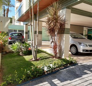 Uma área de estacionamento com dois carros, um edifício com paredes revestidas de azulejos verdes e brancos, e um pequeno jardim com plantas e uma palmeira.