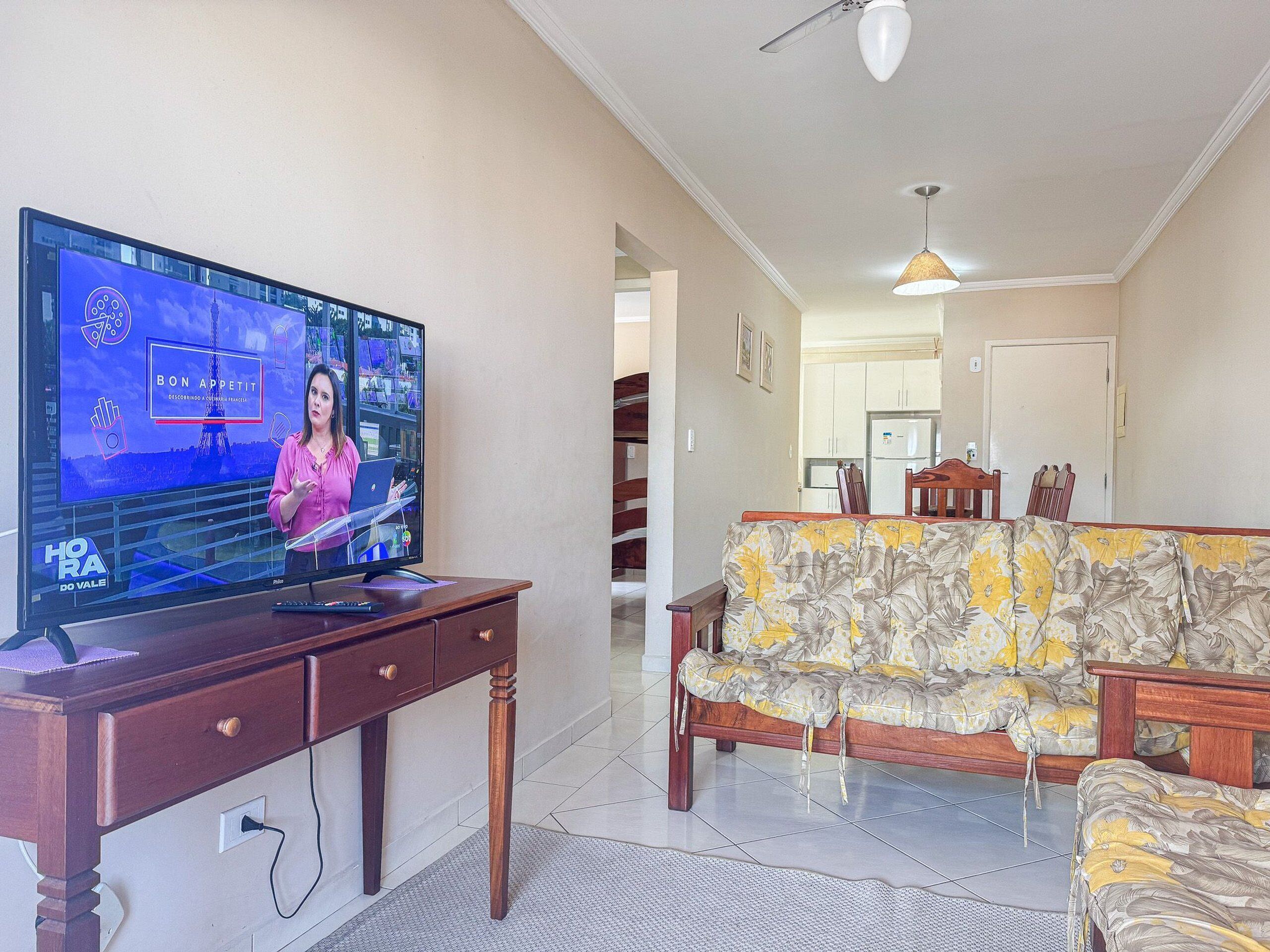 A living room with a TV displaying a news program, a wooden desk, a floral sofa, and a kitchen in the background.