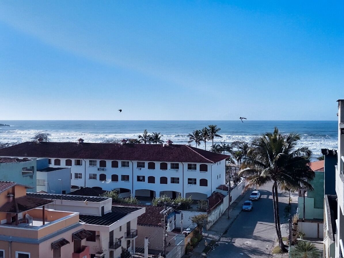 A coastal building with multiple floors, a street with parked cars, and palm trees.