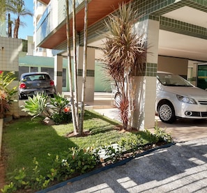 A parking area with two cars, a building with green and white tiled walls, and a small garden with plants and a palm tree.