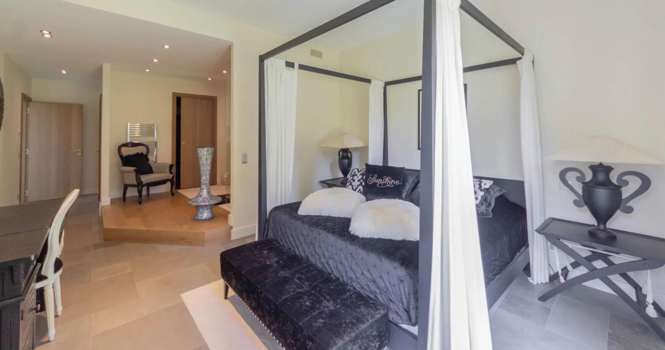 A four-poster canopy bed with a black and white color scheme, a small bedside table with a lamp, and a chair in the adjacent room.