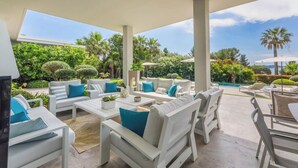 An outdoor patio area with white furniture and blue cushions, surrounded by lush greenery and palm trees. - Superbe Villa vue mer sur st Tropez (Sainte-Maxime)