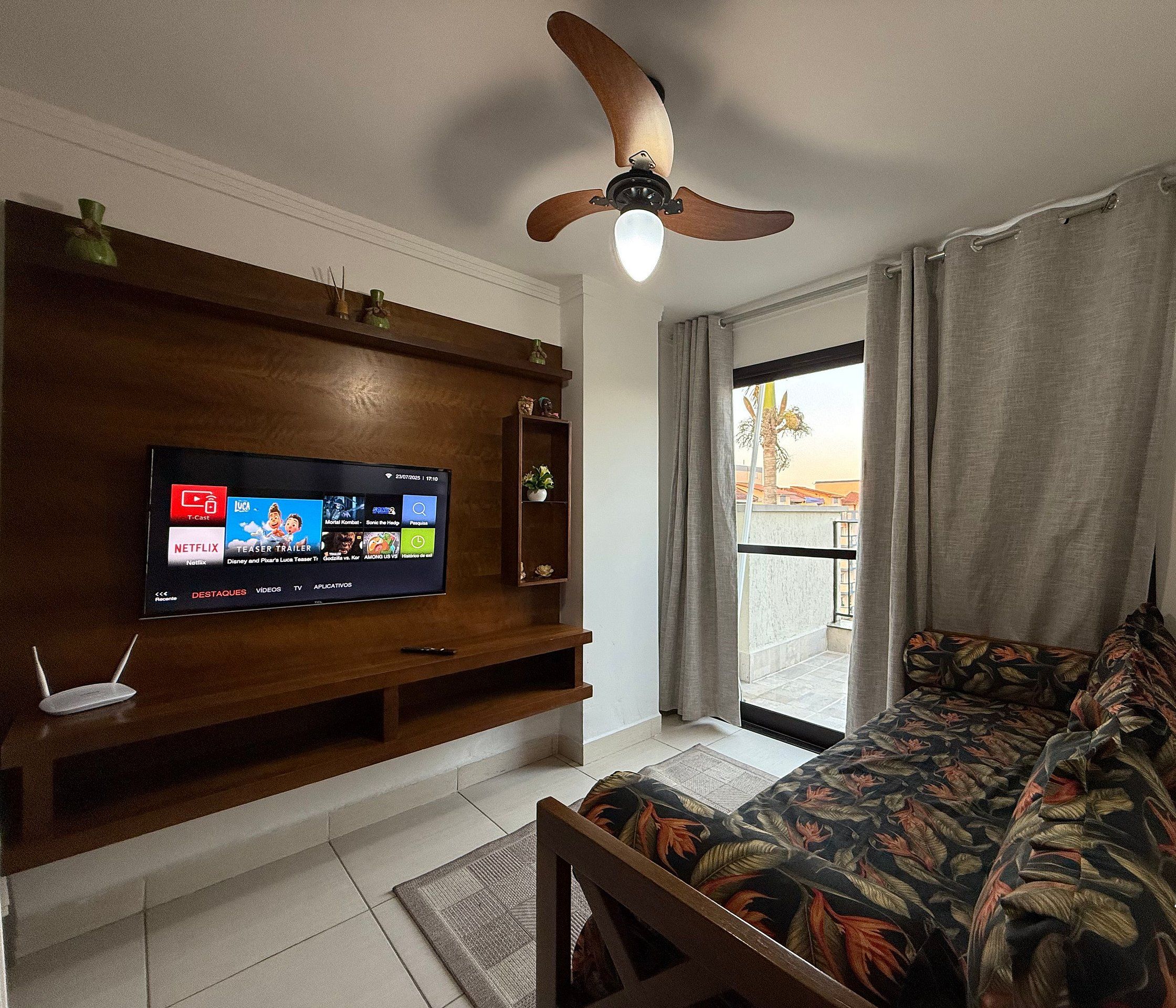 Una sala de estar moderna con un televisor de pantalla plana, mueble de madera para entretenimiento, ventilador de techo y vista al exterior desde un balcón.