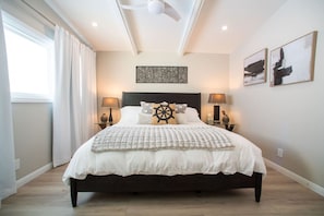 Un dormitorio con cama, dos lámparas de noche, ventilador de techo, ventana con cortinas y obras de arte en las paredes.