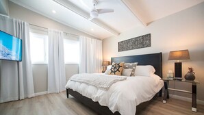 A modern bedroom with a large bed, a ceiling fan, a flat-screen TV, and a bedside table with a lamp. - La Jolla Windansea Luxury Cottage (San Diego)