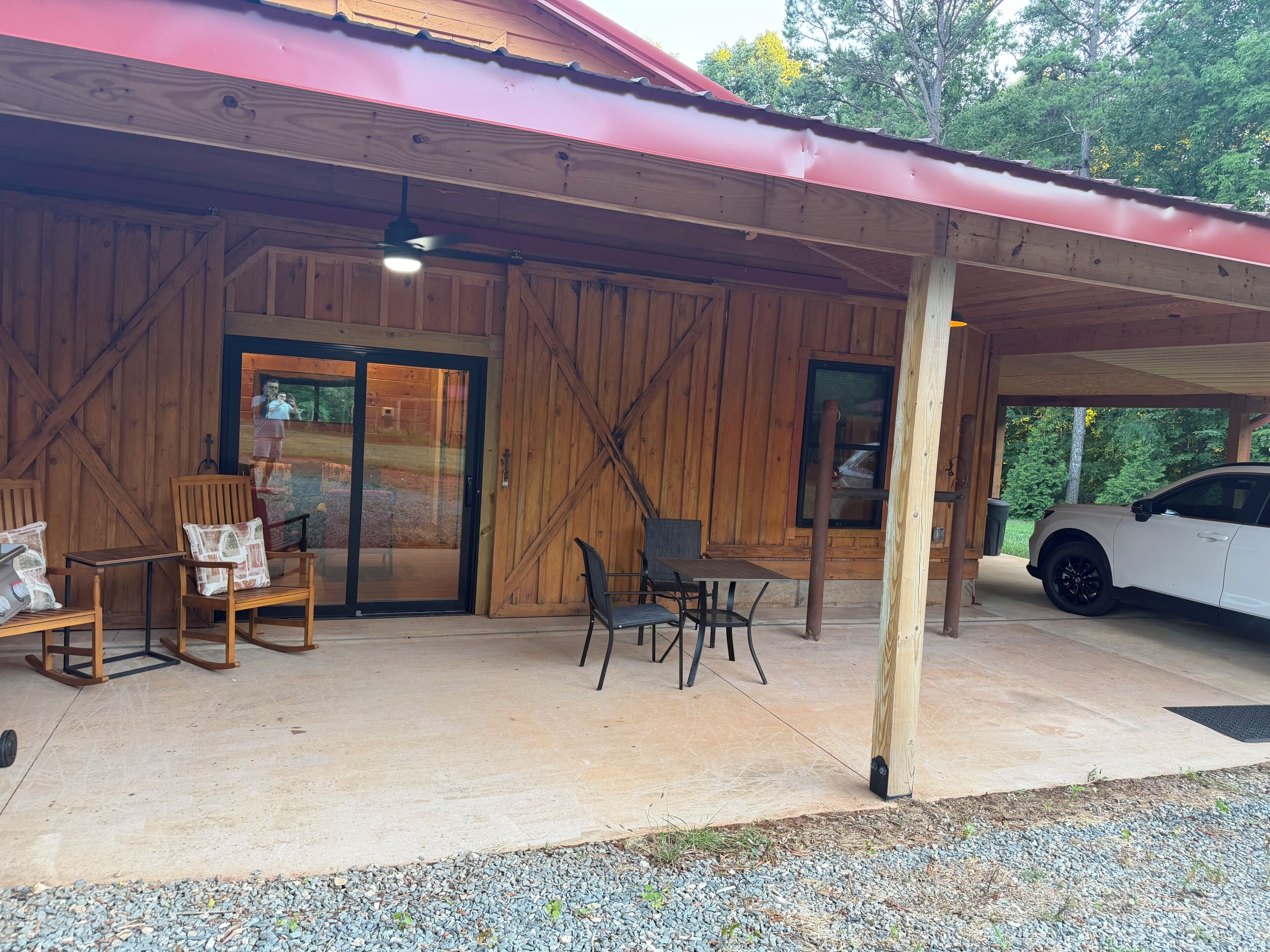A covered outdoor area with a car parked inside, wooden furniture, and a large sliding door.