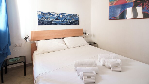 A neatly made bed with white linens, a wooden headboard, and a nightstand with a lamp. There are towels on the bed and a small table with a lamp beside the bed. - Petali di Rosa – A Due Passi dalla Spiaggia (Loiri)