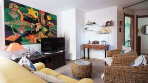 A living room with a large abstract painting, a television, wicker furniture, and a glass coffee table. - Petali di Rosa – A Due Passi dalla Spiaggia (Loiri)