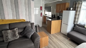 Un salon moderne avec un canapé, une table basse et un coin repas comprenant une table et des chaises. On aperçoit au fond une cuisine équipée d’un réfrigérateur et d’une cuisinière.