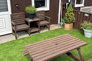 Un porche de madera con una mesa y sillas, una planta en maceta y un pequeño jardín con césped artificial.