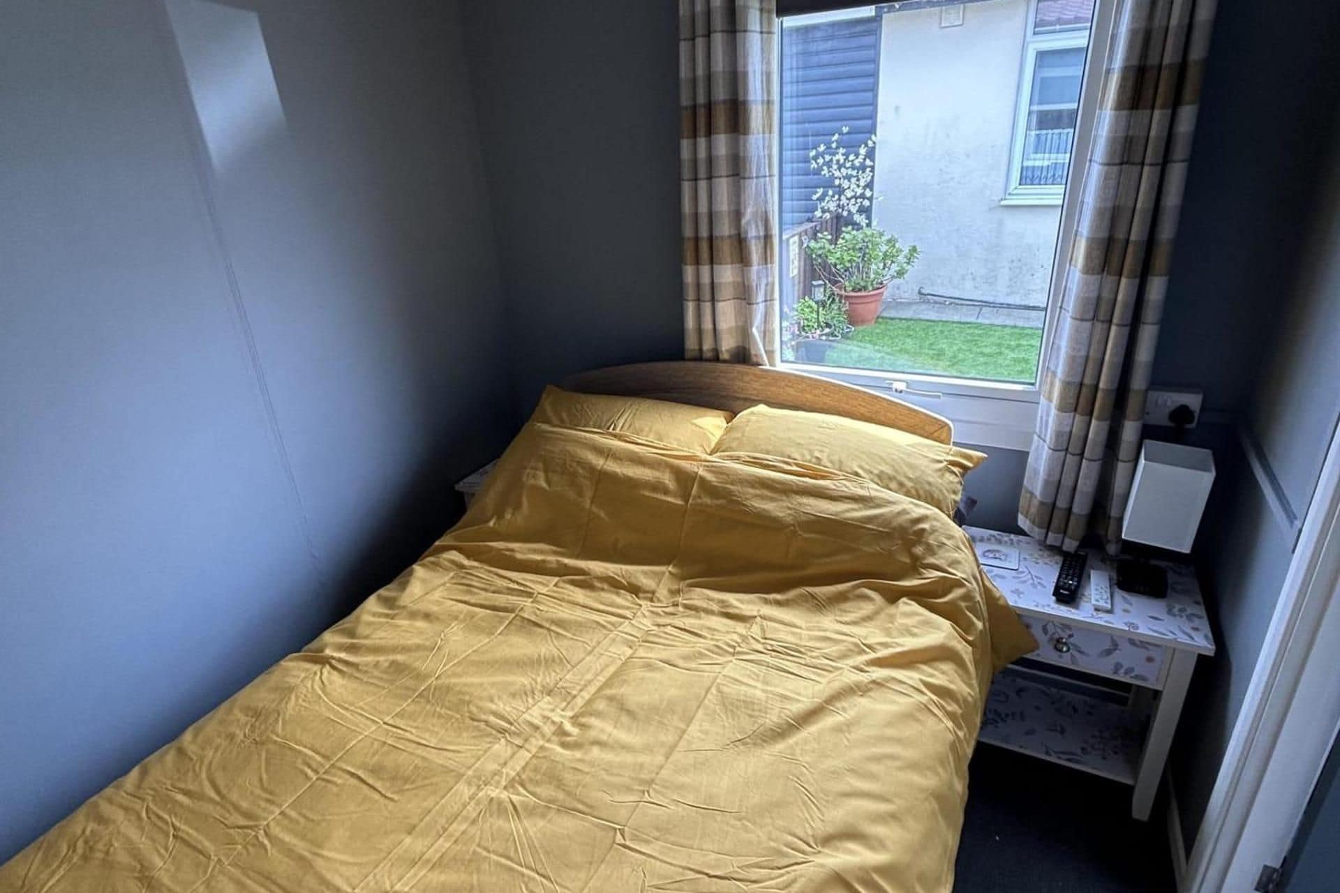 A small, single-bed room with a window, a bedside table, and a view of the outside.