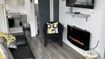 A living room with a black leather sofa, a black armchair with a patterned cushion, a flat-screen TV mounted on the wall, and a fireplace with logs.