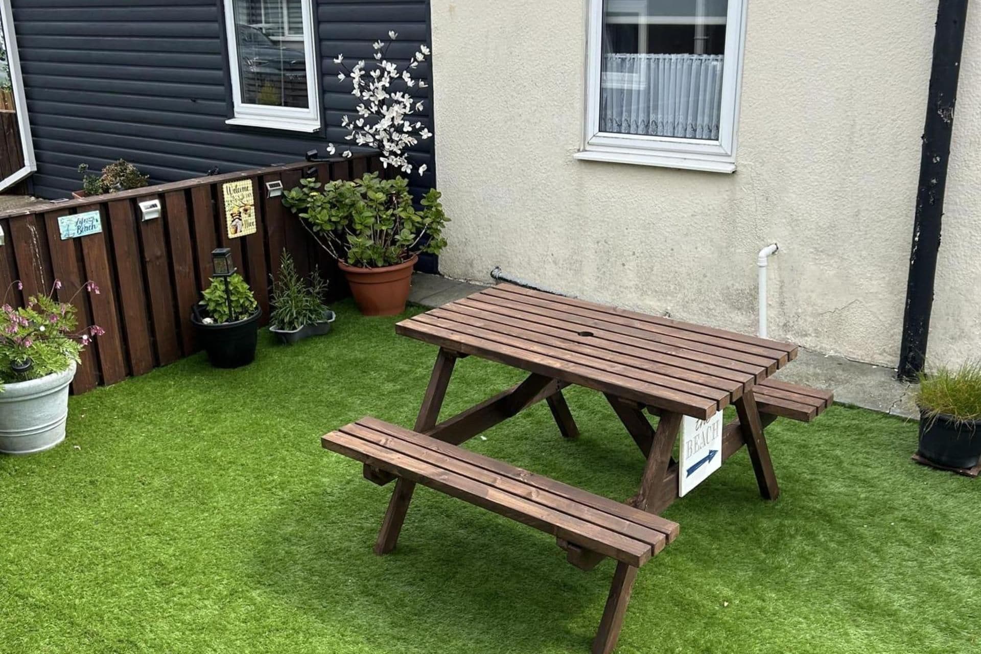 Une table de pique-nique en bois, avec des bancs, située dans un espace jardin agrémenté de gazon synthétique, entourée de plantes en pot et d’une clôture en bois.