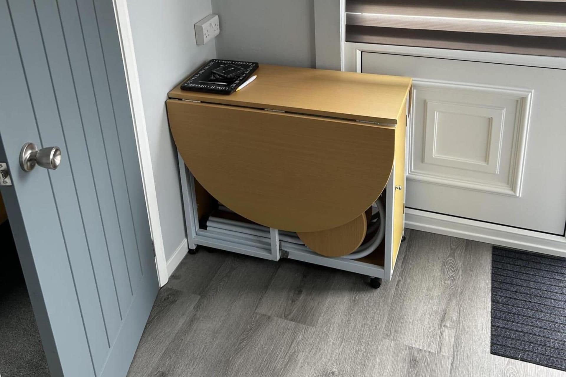 A compact, wall-mounted storage unit with a pull-out drawer, positioned in a corner of a room with a door and a cabinet.