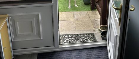 Een kamer met een grijze deur, een klein tapijt en een hond die buiten staat op een gazon met potplanten.