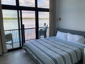 A hotel room with a large window offering a view of a lake and trees. The room includes a bed with a patterned bedspread, a chair, and a small table.