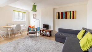 Een woonkamer met een grijze bank, een witte eettafel met stoelen, een televisie op een houten dressoir en een kleurrijk abstract schilderij aan de muur.