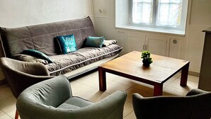 Una sala de estar con un sofá, dos sillones, una mesa de centro, una ventana con persianas y un cuadro de empotrado eléctrico en la pared.