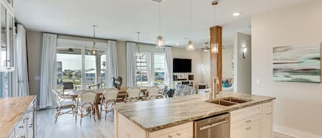 A spacious kitchen with a large island, wooden cabinets, and a dishwasher. There is a dining area with wicker chairs and a table, and a large window providing a view of the outside.