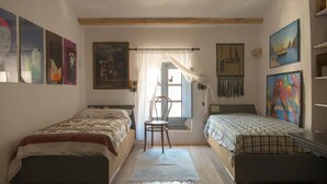 A bedroom with two single beds, a chair, a window with curtains, and various wall art. - Sardinia RE - Alma Boh Mien Stunning View (Alghero)