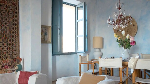 A room with a white sofa, a dining table with chairs, a chandelier, and a window with blue shutters. - Sardinia RE - Alma Boh Mien Stunning View (Alghero)