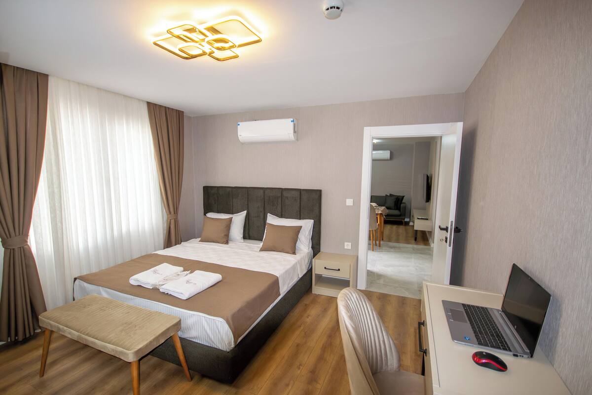 A hotel room with a bed, bedside table, chair, desk with a laptop, and an air conditioning unit.