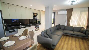 Uma sala de estar moderna com um sofá modular cinza, uma mesa de jantar posta com pratos e talheres, e uma área de cozinha com uma televisão de tela plana.
