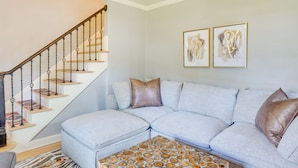 A living room with a grey sectional sofa, a patterned ottoman, a staircase with wooden steps and metal railings, and two framed abstract artworks on the wall. - Walkable Thiensville Retreat Near Hiking! (Mequon)