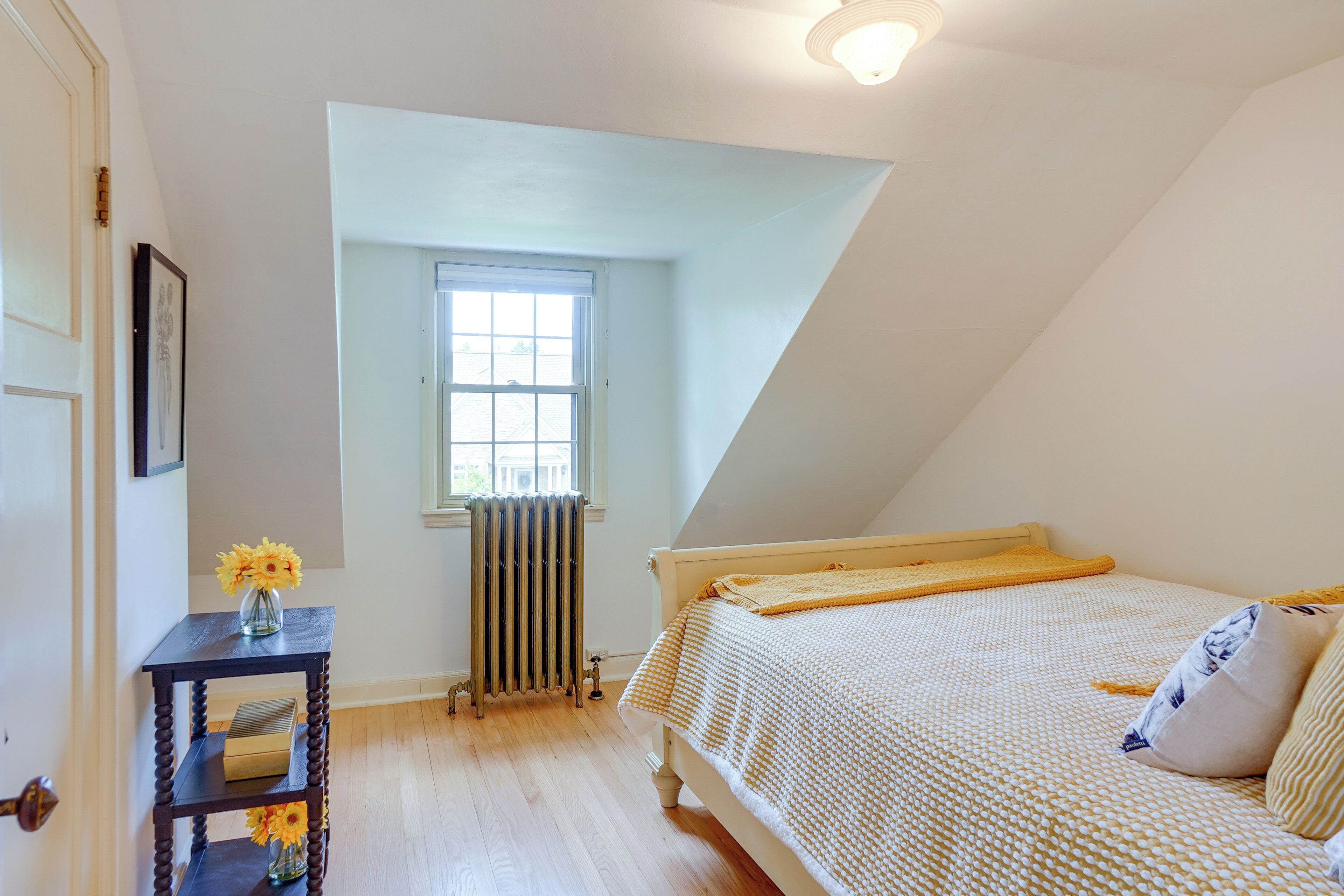 A bedroom with a sloped ceiling, a window, a radiator, a bedside table with a vase of flowers, and a bed with a patterned bedspread.