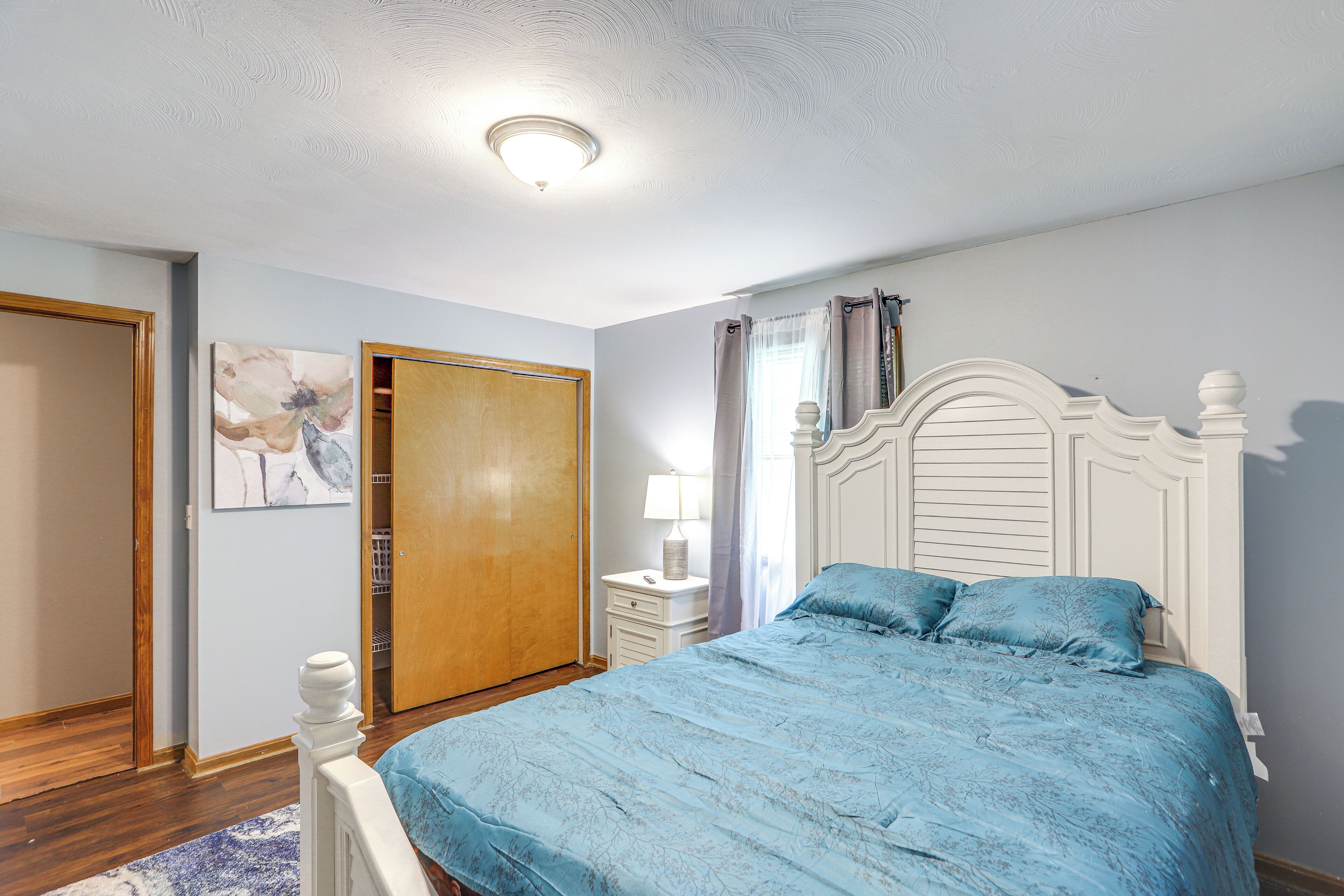 A bedroom with a bed, a nightstand, a lamp, a door, and a wall with a painting.