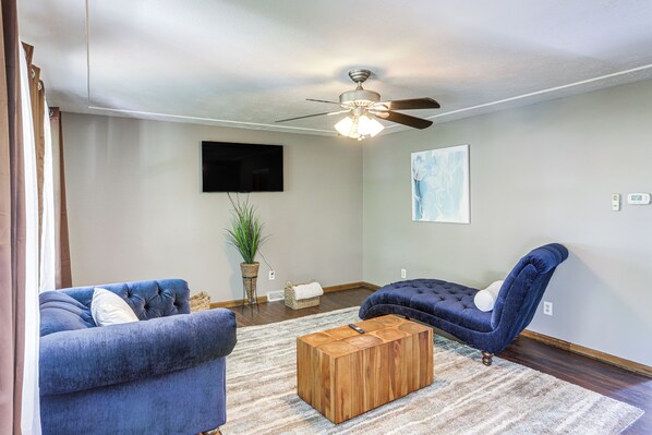 A living room with a ceiling fan, a large flat-screen TV, a blue sofa, a blue armchair, and a wooden coffee table. - Lake Erie Family Getaway in Amherst: Pets Welcome! (Amherst)