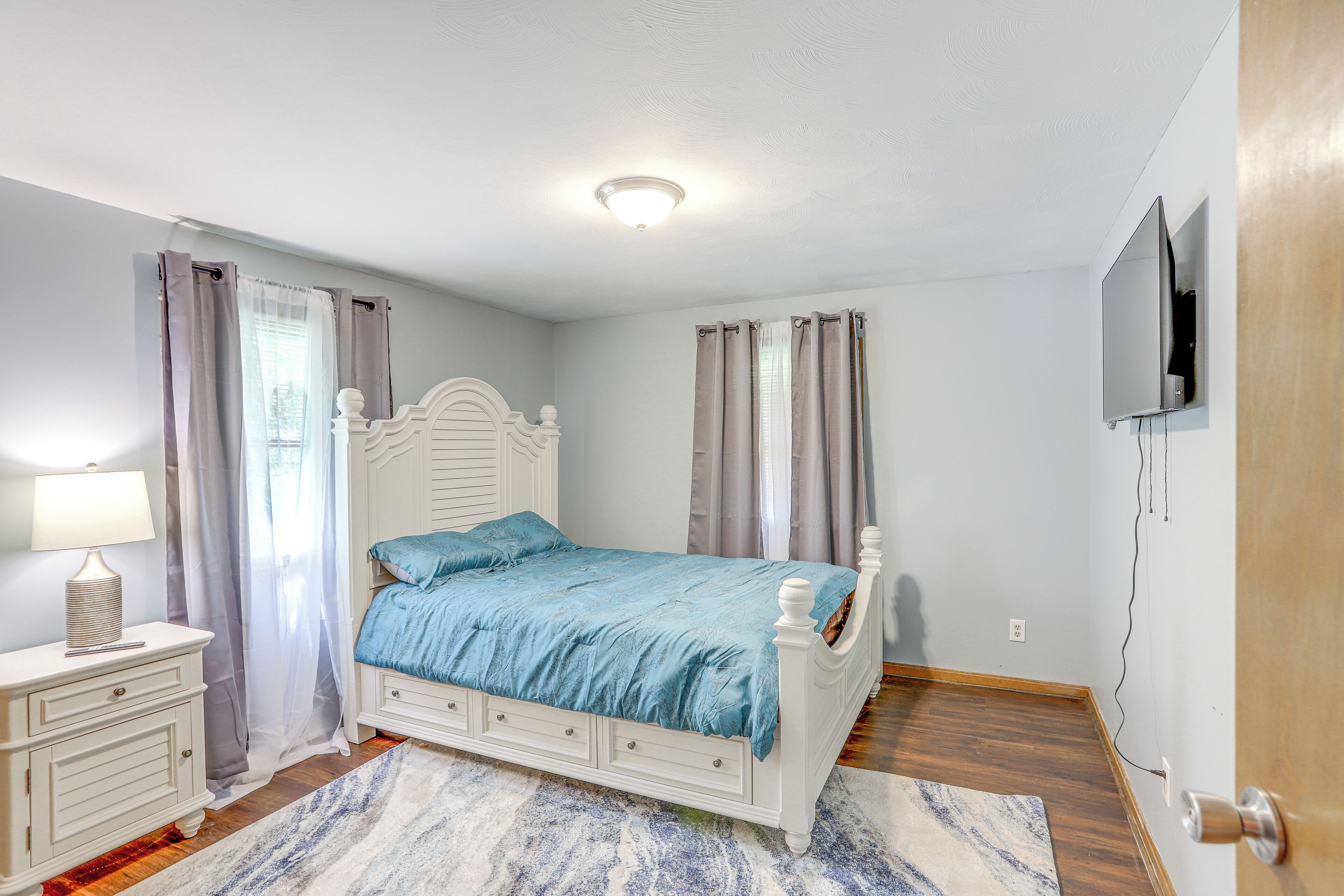 Une chambre avec un lit blanc, une table de chevet avec une lampe, un téléviseur fixé au mur et un plancher en bois.