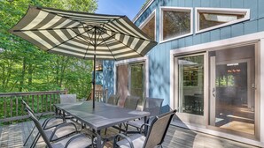 A deck with a table and chairs, a striped umbrella, and a house with large windows and a door.