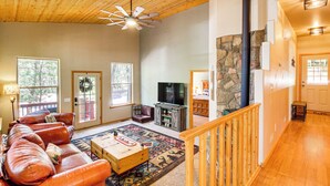 A spacious living room with a wooden ceiling, a stone fireplace, a large window, and a television. - Walk to Rainbow Lake: Peaceful Cabin w/ Fire Pit (Lakeside)